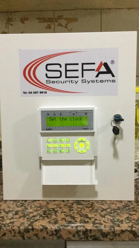 SEFA Security Systems alarm control panel with green LCD display, keypad, and key lock mounted on a white metal box.