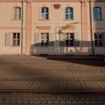View of a historic hospital building facade with tram shadows cast on the pavement.